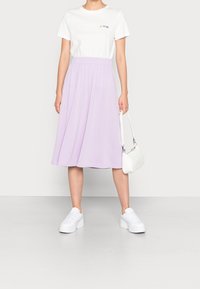 Witte t-shirt met meerkleurige print, gecombineerd met een lavendelkleurige midi-jurk. Model draagt witte sneakers en heeft een kleine witte handtas.