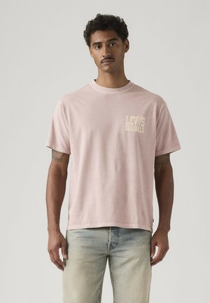 Mężczyzna o krótkich kręconych włosach i wąsach, ubrany w jasnoróżową koszulkę Levi's Originals i wyblakłe jasnoniebieskie dżinsy, stojący na jednolitym tle.