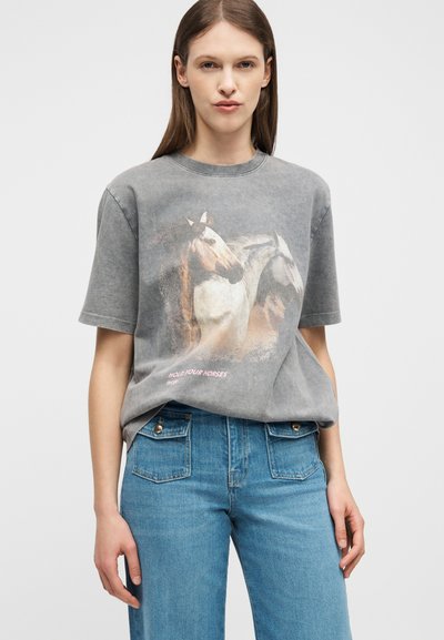 Νεαρή γυναίκα που φορά ένα ξεβαμμένο γκρι T-shirt με γραφικό δύο αλόγων και το κείμενο "Κράτα τα άλογά σου", συνδυασμένο με μπλε τζιν.