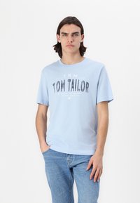 Světle modré bavlněné tričko s výrazným černým grafickým potiskem, který zní "TOM TAILOR." Krátké rukávy a design s kulatým výstřihem.