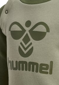 Zielona bluza z dużym oliwkowym logo o abstrakcyjnym wzorze oraz nazwą marki "hummel" nadrukowaną wyraźnymi literami. Widoczna tekstura tkaniny.