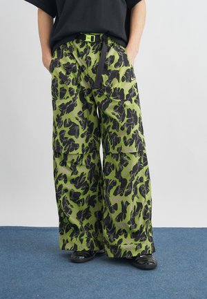 Osoba ubrana w zielone spodnie cargo o szerokich nogawkach z czarnym abstrakcyjnym wzorem, czarny pasek, czarne buty i czarny top, stojąca na niebieskim dywanie.