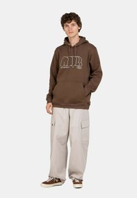 Brauner Hoodie mit weißem Grafikdesign, kombiniert mit hellgrauen Cargo-Hosen mit Taschen. Das Modell trägt braune Sneakers.