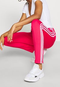 Růžové sportovní kalhoty s bílými bočními pruhy, vyrobené z hladkého materiálu. Nosí se s bílým upnutým trikem a bílými teniskami. Jednoduchý design.