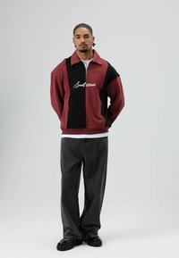 Sweat-shirt bicolore bordeaux et noir avec un col à zip partiel, orné de texte blanc en cursive. Porté avec un jean gris foncé et des chaussures noires.