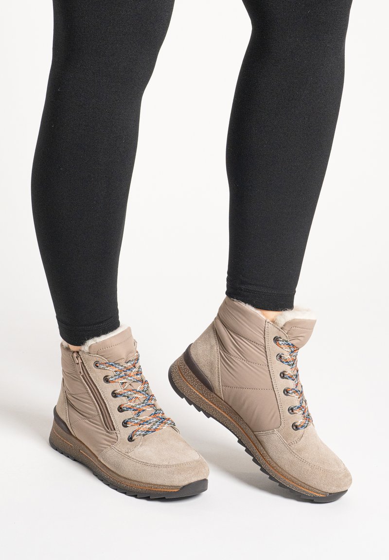 Beige Winter-Stiefeletten mit bunten Schnürsenkeln, seitlichen Reißverschlüssen und Fellfutter, getragen zu schwarzen Leggings vor neutralem Hintergrund.