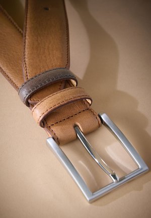 Nærbillede af et lysebrunt læderbælte med syninger langs kanten og en sølvfarvet rektangulær metalspænde på en beige overflade.