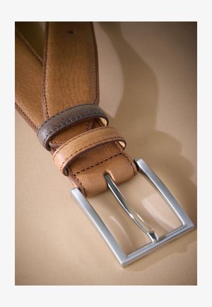 Nærbillede af et lysebrunt læderbælte med syninger langs kanten og en sølvfarvet rektangulær metalspænde på en beige overflade.