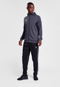 Sportig träningsdräkt bestående av en mörkgrå zip-up huvtröja och svarta byxor med en chevronmönster längs sidorna, tillverkad i ett mjukt tyg.