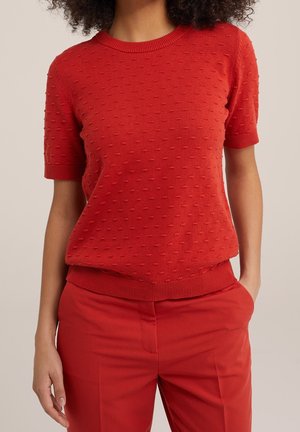 Woman wearing a textured, short-sleeve red knit top and matching red pants, standing with one hand in pocket against a plain background.