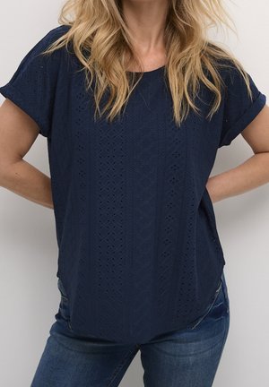 Haut à manches courtes bleu marine en tissu texturé avec motifs en losange. Présente une coupe ample et un ourlet arrondi, associé à un jean en denim.