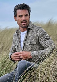 Braunes Karo-Flanellhemd mit zwei Brusttaschen, getragen über einem weißen T-Shirt. Graue Hose, sitzt im hohen Gras unter einem bewölkten Himmel.