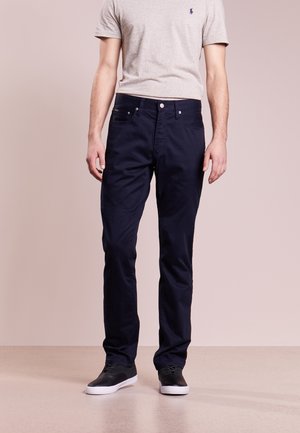 Mężczyzna ubrany w granatowe spodnie, beżową koszulę z krótkim rękawem oraz czarne trampki z białą podeszwą stojący na jednolitym tle.