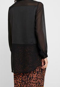 Femme portant un blouse noire transparente à manches longues sur une jupe en imprimé léopard avec des nuances d'orange, de marron et de noir, posant avec la main détendue.