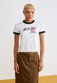 Witte cropped T-shirt met zwarte ringerhalslijn en mouwen, met de tekst "Ik ben er dan" en cartoonafbeeldingen. Gecombineerd met een bruine rok.