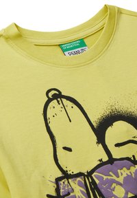 Žluté tričko s černou a fialovou grafikou Snoopovy tváře, se zelenou nášivkou United Colors of Benetton a Peanuts uvnitř límce.
