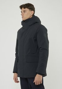 Giacca puffer da uomo blu navy con cappuccio staccabile, grandi tasche frontali e polsini elasticizzati. Realizzata in tessuto liscio e resistente agli agenti atmosferici.