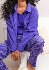 Fialová fleecová bunda s knoflíkovými zapínáními přes sladěný fialový top a kalhoty, zdobená bílými tečkovanými vzory ve vlnitých liniích.