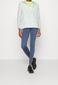 Veste à demi-zip vert clair avec capuche, accents réfléchissants, associée à des leggings bleus et des chaussures de sport noires. Textures lisses et extensibles.