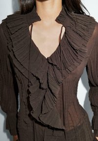 Blouse en chiffon marron avec des volants plissés le long de l'encolure et du buste ; présente un décolleté en V profond et des manches longues. Tissu texturé dans l'ensemble.
