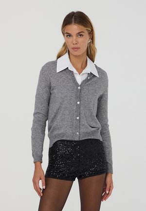 Cardigan grigio lavorato a maglia con scollatura a V, con bottoni sul davanti, indossato sopra una camicia bianca con colletto. Abbinato a pantaloni corti neri con paillettes.