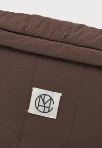 Καφέ ραμμένο ύφασμα με μια λευκή ορθογώνια ετικέτα που έχει ένα μαύρο κυκλικό λογότυπο με στυλιζαρισμένα διασυνδεδεμένα γράμματα.