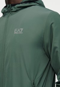 Κοντινό πλάνο ατόμου που φοράει πράσινο μπουφάν με φερμουάρ και κουκούλα, με το λογότυπο EA7 Emporio Armani στο στήθος και τσέπη με φερμουάρ στο πλάι.