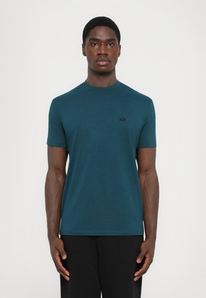 Man wearing a teal short-sleeve crew neck t-shirt with black A|X logo on the chest, standing against a plain white background.