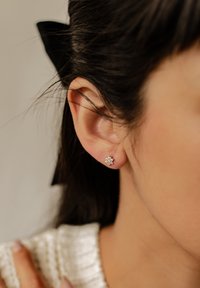 Aretes de plata en forma de flor, con múltiples pequeñas piedras preciosas transparentes. Un lazo negro es visible en el cabello.