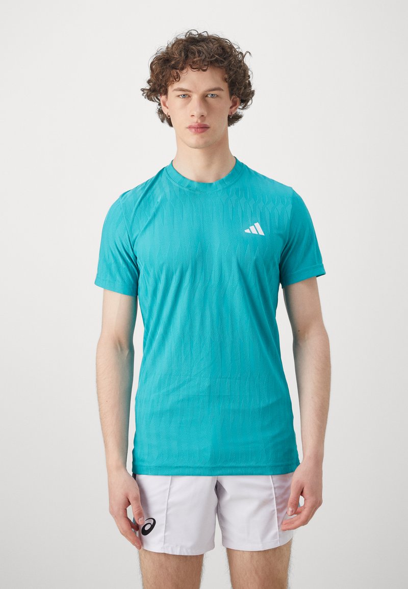 Türkiz színű sport póló könnyű, texturált anyagból; rövid ujjakkal és kerek nyakkal rendelkezik; a mellkasán egy fehér logó részlet található.