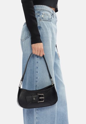 Mujer con vaqueros azul claro y top corto de manga larga negro sosteniendo un pequeño bolso negro con detalle de hebilla plateada.