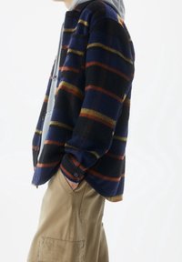 Camicia in pile con motivo a quadri blu, neri e arancioni, dotata di chiusura con bottoni e vestibilità comoda, indossata sopra un maglione grigio.