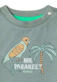 T-shirt verte pour enfants avec un perruche brun et beige, un palmier, une plante feuillue, et le texte "MR. PARAKEET NOPPIES" sur le devant.