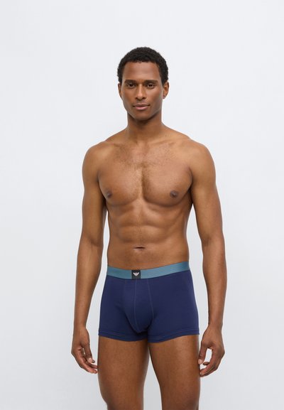 Tamsiai mėlyni bokseriai su minkštu audiniu, kontrastinga žalsvai mėlyna juostos juosta ir juodu logotipo lopu priekyje. Aptempta forma pabrėžia kūno linijas.