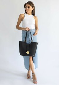 Czarna skórzana torba tote o wydłużonym kształcie, złotych akcentach i centralnym pasku. Niedokończona dolna krawędź, gładka tekstura.