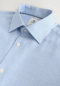 Light blue textured dress shirt with white buttons and a pointed collar laid flat on a white surface.