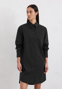 Zwart blousejurk met een kraag, lange mouwen en een relaxte pasvorm. Subtiele textuur en gladde stof. De zoom zit boven de knie.