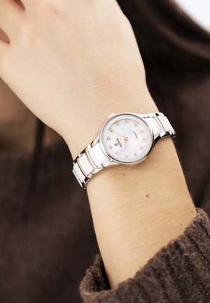 Poignet d'une femme portant une montre bracelet en métal argenté avec un cadran blanc et de petits marqueurs en cristal, sur un fond de pull marron.