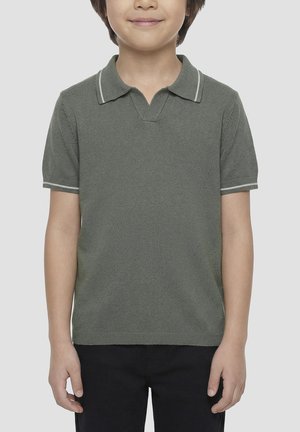 Dijete nosi kratku olivno zelenu pletenu polo majicu s bijelim obrubom na ovratniku i rukavima, uparenu s crnim hlačama, stoji ispred jednobojne pozadine.