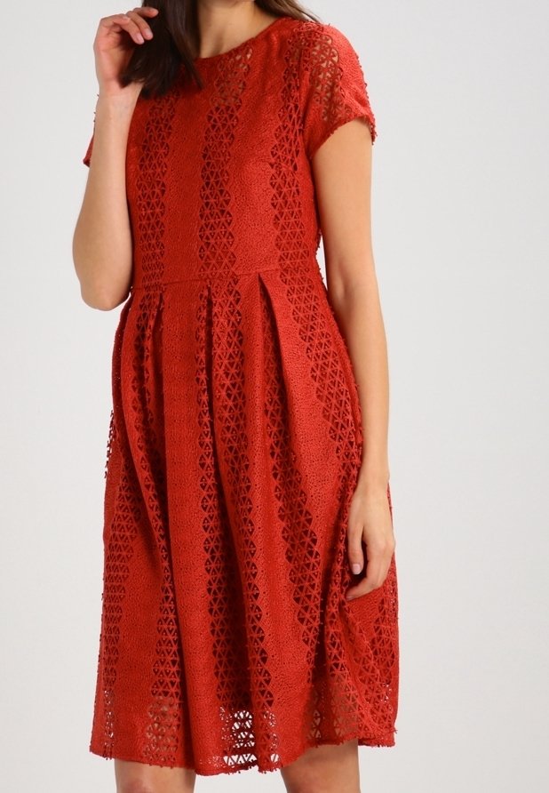 Femme portant une robe rouge en dentelle au genou, à manches courtes, avec des motifs géométriques découpés, debout devant un fond uni.