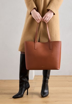 Manos sosteniendo un bolso tote de cuero marrón sobre botas negras de tacón hasta la rodilla y un abrigo camel sobre un fondo de suelo de madera clara.