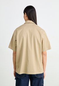 Lys kortermet skjorte i beige laget av lett stoff, med krage og synlige sømmer, sammen med mørkeblå bukser.