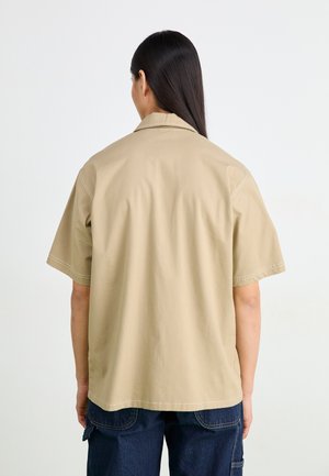 Camisa de manga corta de color beige, hecha de un tejido ligero, con cuello y detalles de costura visibles, usada con pantalones azul oscuro.