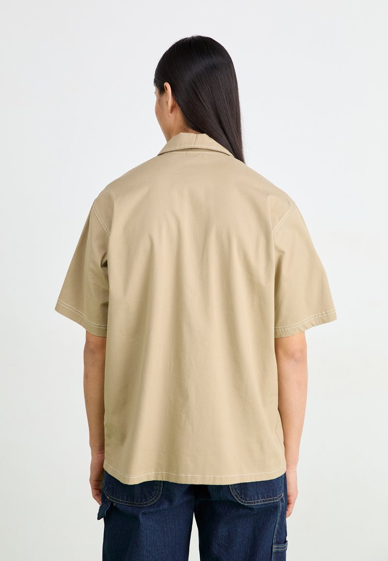 Lys kortermet skjorte i beige laget av lett stoff, med krage og synlige sømmer, sammen med mørkeblå bukser.