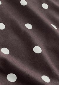 Dunkelbrauner Stoff mit gleichmäßig verteilten großen weißen Tupfen, der weiche Falten und Texturdetails zeigt.