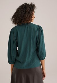 Türause Bluse aus strukturiertem Stoff, mit Puffärmeln und gerafftem Ausschnitt, kombiniert mit einem dunkelbraunen Rock. Sauberes, minimalistisches Design.