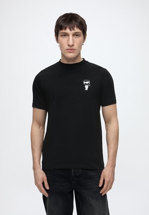 Jeune homme portant un t-shirt noir avec un petit graphique blanc représentant un astronaute sur le côté gauche de la poitrine, debout devant un fond clair uni.