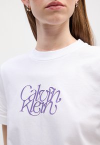 T-shirt en coton blanc avec un logo "Calvin Klein" en script violet sur la poitrine, col rond et manches courtes.