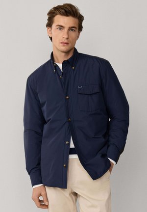 Joven con cabello castaño claro, vestido con una chaqueta azul marino abotonada sobre una camisa blanca y pantalones beige, de pie con una mano en el bolsillo.