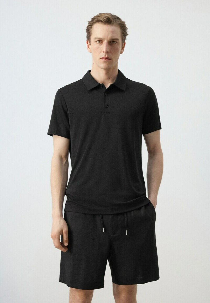 Homme debout face à l'avant, portant un polo noir à manches courtes et un short noir à cordon, sur un fond uni clair.